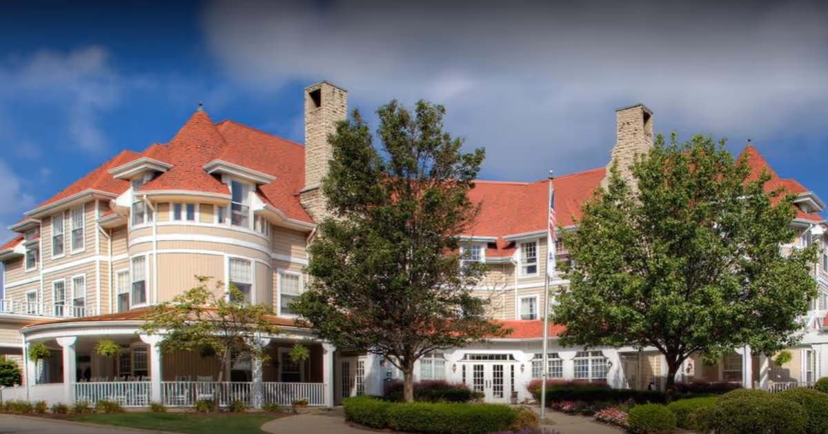 Exterior view of a large senior living facility building with beige siding and a red roof. The building features multiple windows, two prominent chimneys, and a covered porch area. There are several trees and well-maintained bushes and flowers in front of the building under a partly cloudy sky.