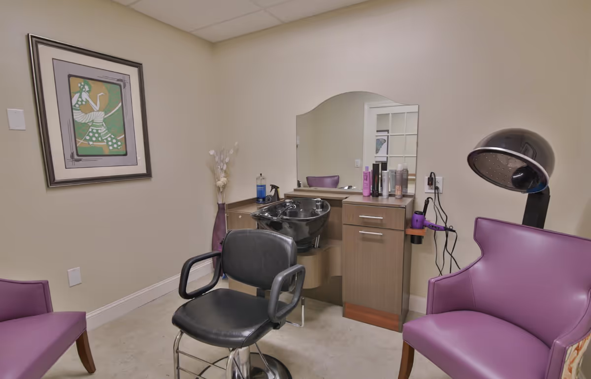 A small salon area in a senior living facility with a black salon chair in front of a hair washing station, a large mirror, and a cabinet with hair care products. There are two purple armchairs, a hair dryer hood, a framed artwork on the wall, and a tall vase with decorative branches in the corner.