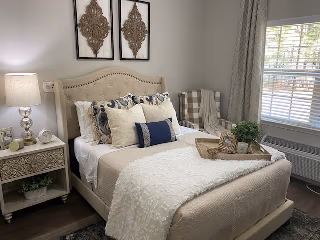 A cozy bedroom with a beige upholstered bed adorned with multiple decorative pillows in shades of cream, navy, and patterned designs. A soft textured white throw blanket is draped over the bed. Next to the bed is a nightstand with a decorative lamp, a small clock, and framed photos. Two ornate wall art pieces hang above the headboard. A window with blinds and light gray curtains lets in natural light, and a checkered armchair with a throw blanket is positioned in the corner near an air conditioning unit.