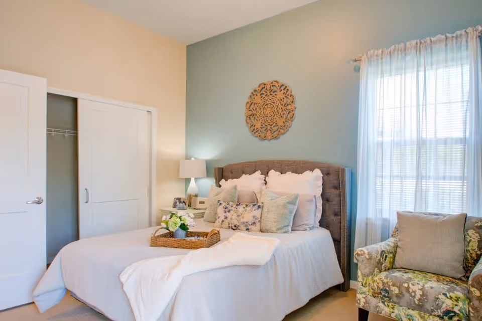A cozy bedroom with a neatly made bed featuring multiple pillows and a white blanket. A decorative wooden wall piece hangs above the tufted headboard. To the right, a window with sheer white curtains allows natural light to fill the room. A floral-patterned armchair with a beige cushion is positioned near the window. On the left side, there is a nightstand with a lamp, a photo frame, and a small clock. A closet with sliding doors is partially open.