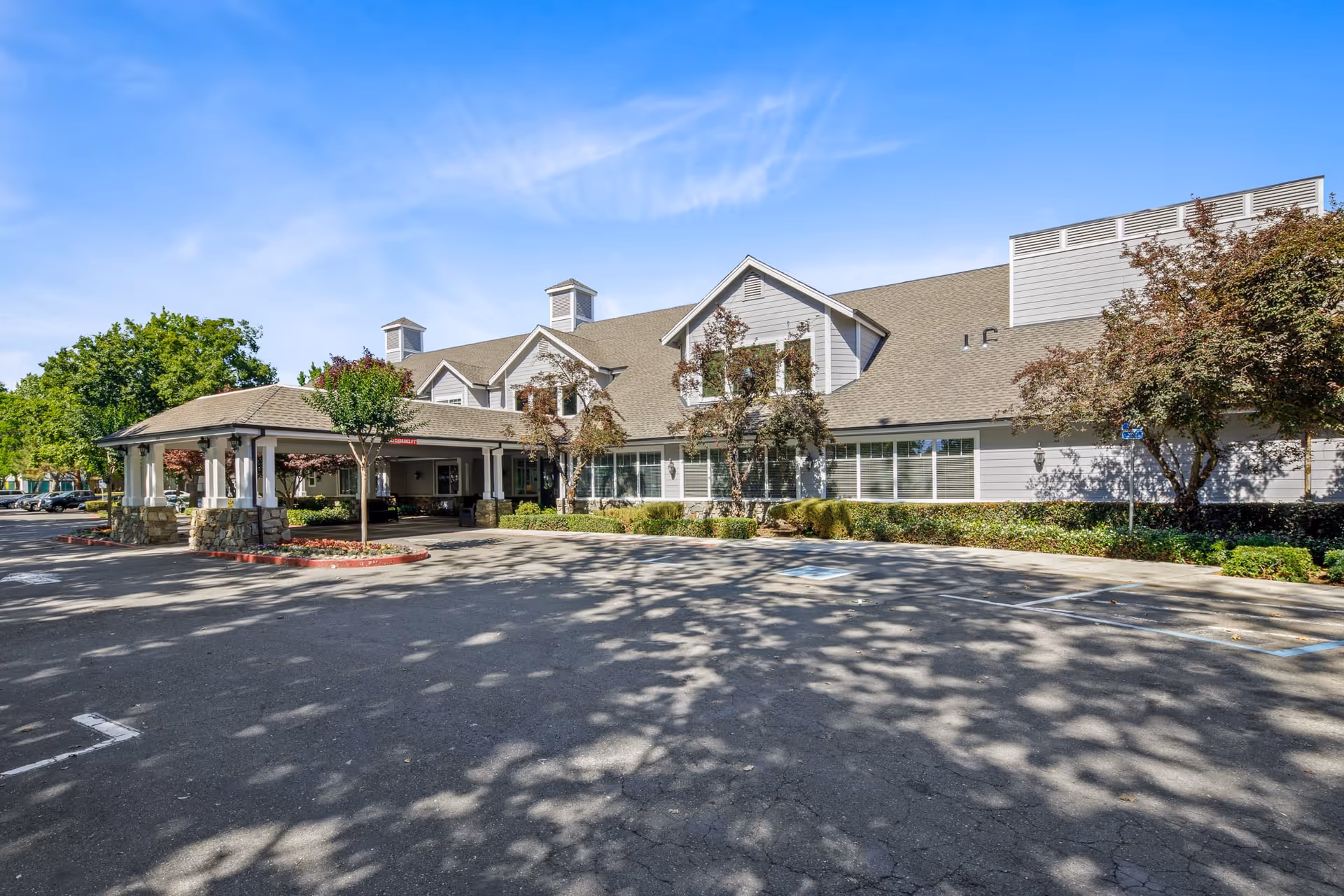 Exterior view of Oakmont Of Brookside senior living facility showing a large building with a covered entrance, surrounded by trees and a parking lot under a clear blue sky.