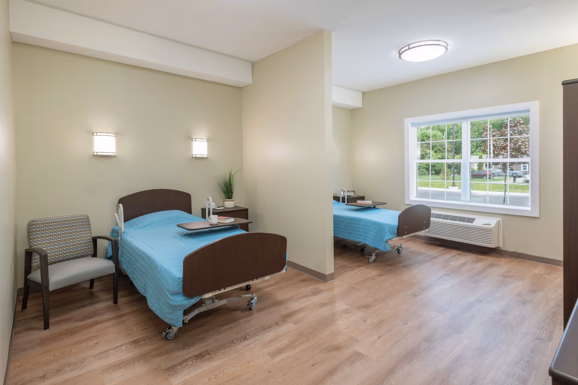 A shared patient room in a healthcare facility with two hospital beds separated by a partial wall. Each bed has a blue blanket and a bedside table with a plant and some books. There is a chair next to one bed and a large window letting in natural light. The room has light-colored walls and wood flooring.