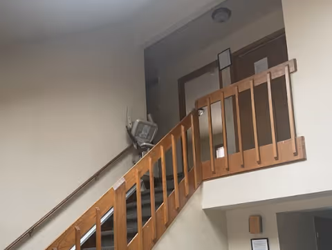 Interior view of a stairway with wooden railings leading to a hallway with doors. The walls are painted light beige and there is a handrail on the left side of the stairs. A stair lift is installed on the right side of the staircase.