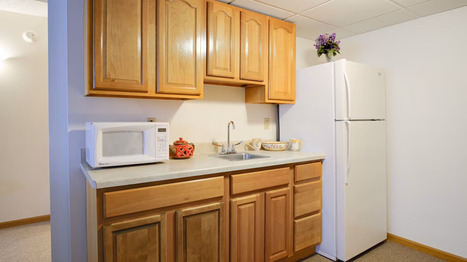 A small kitchen area with wooden cabinets, a white microwave on the countertop, a small sink with a faucet, a white refrigerator, and a decorative flower arrangement on top of the refrigerator. There are also some cups and a bowl on the counter.