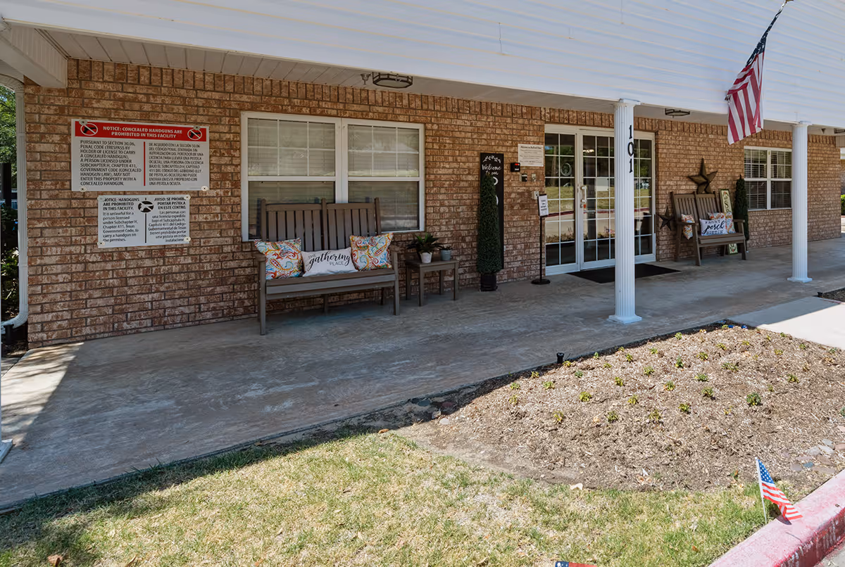 Front porch area of a brick building with two wooden benches adorned with decorative pillows, small potted plants on a side table, an American flag hanging from a white column, and multiple signs including a notice about concealed handguns. The entrance features glass double doors and the building number 101 is visible on a column.