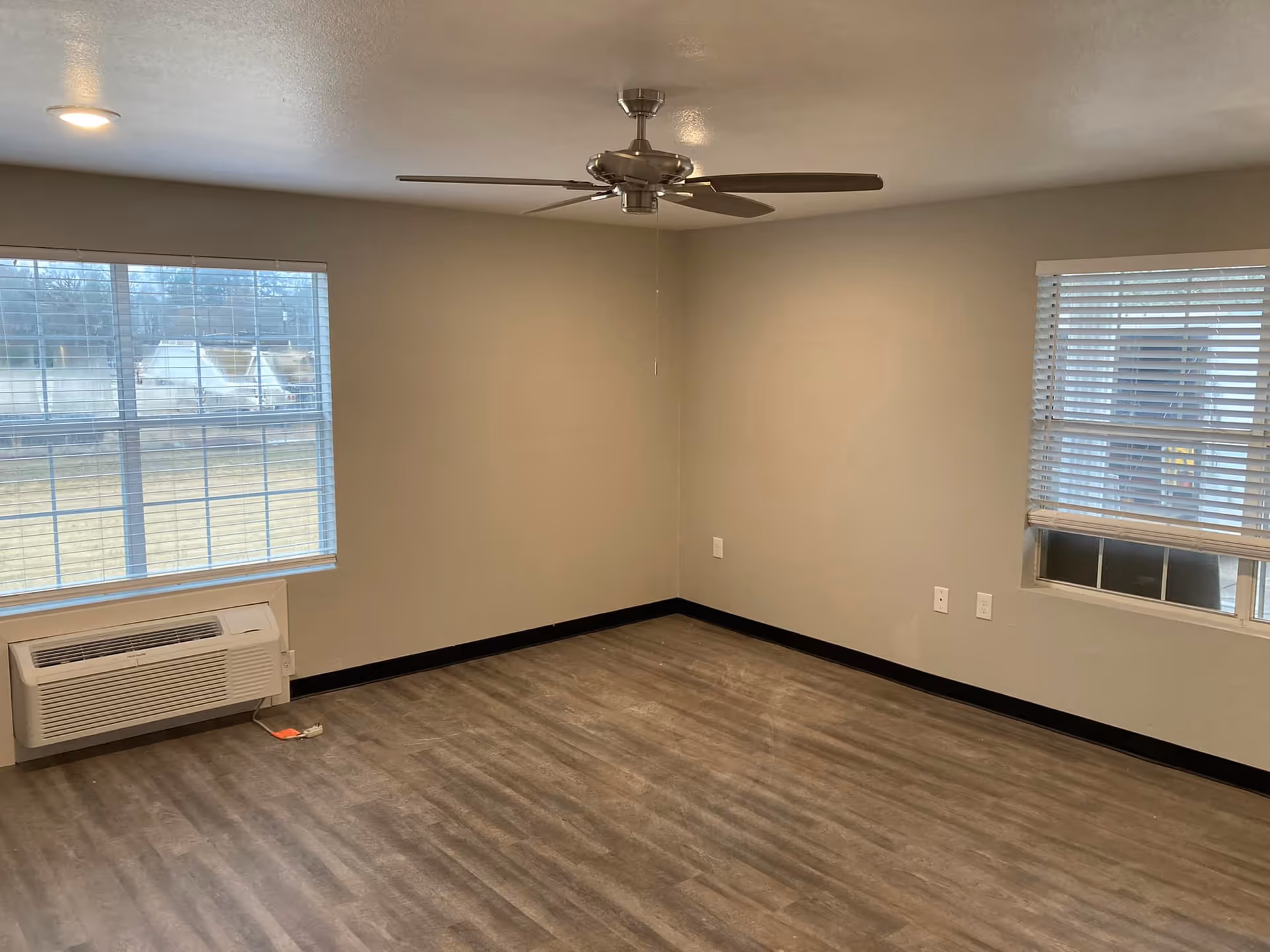 Empty room with two windows with blinds, a ceiling fan with light, wood-style flooring, beige walls, and a wall-mounted air conditioning unit below one window.