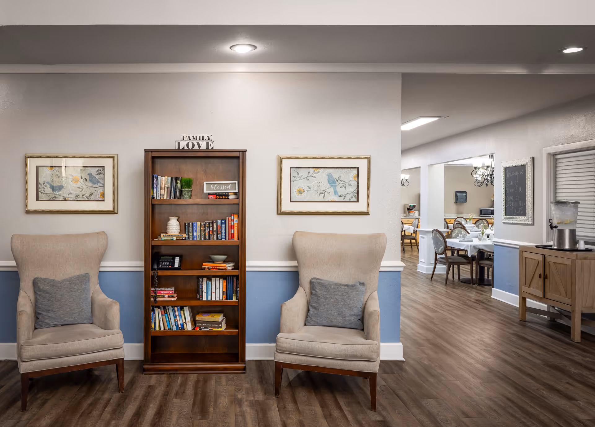 A cozy interior space featuring two beige armchairs with gray cushions on either side of a wooden bookshelf filled with books and decorative items. Above the bookshelf is a sign that reads 'FAMILY LOVE'. The walls are painted white with a blue lower half, and there are framed bird-themed artworks on either side of the bookshelf. In the background, a dining area with tables and chairs is visible, along with a wooden sideboard holding a water dispenser and cups.