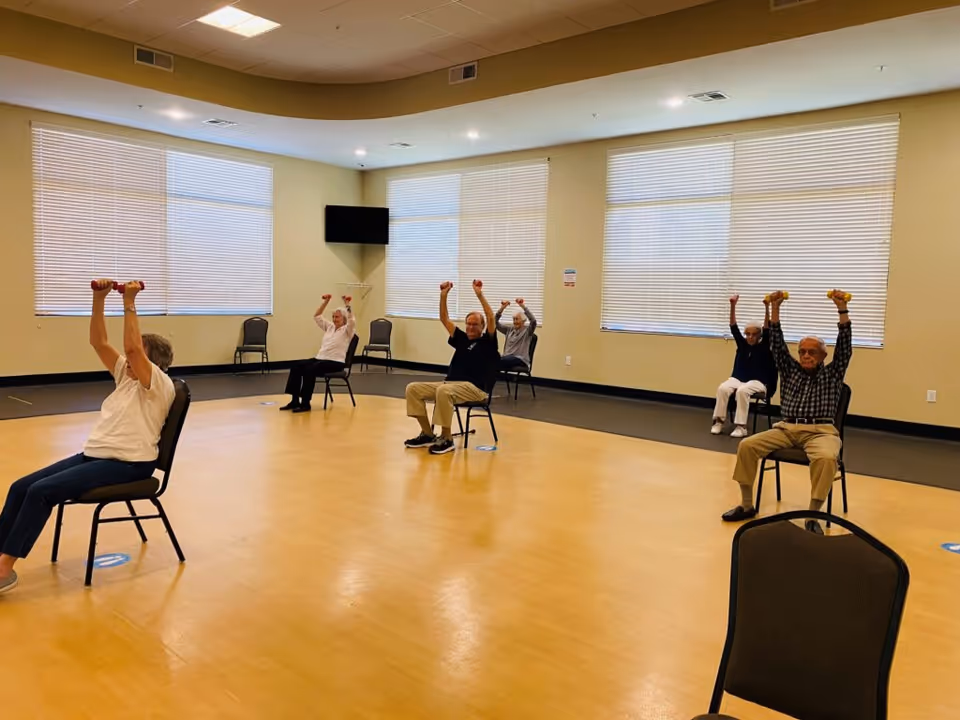 A group of elderly individuals seated on chairs in a spacious room with large windows and blinds, participating in a seated exercise class by lifting small dumbbells above their heads.