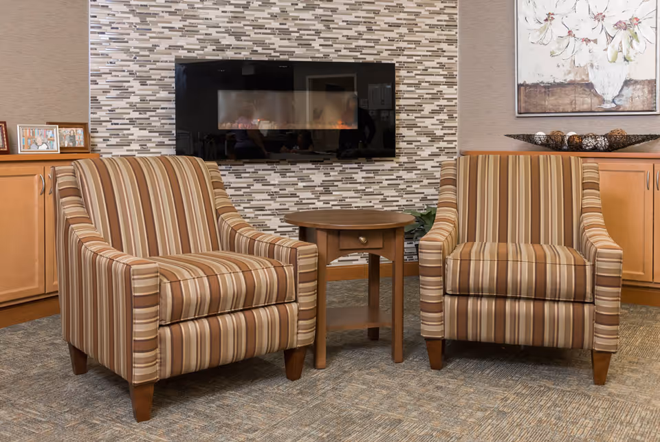 Two striped armchairs flanking a small wooden side table in a lounge area with a wall-mounted electric fireplace and cabinets.