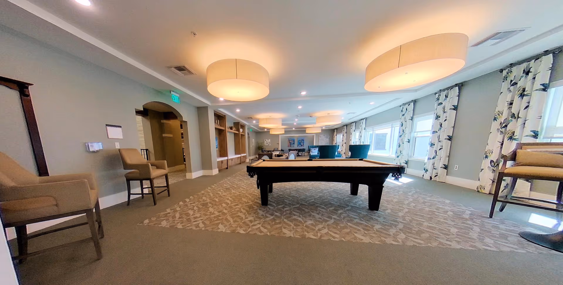 A spacious, well-lit common area with a pool table in the center, surrounded by chairs and large windows with floral curtains allowing natural light to fill the room. The ceiling has multiple large round light fixtures, and the walls are painted a soft gray color.