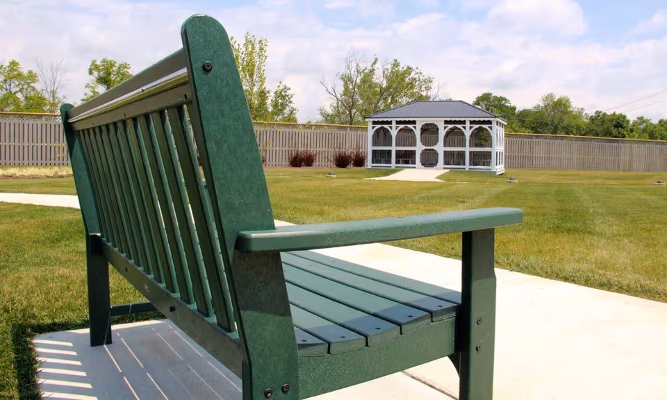 Green wooden bench on a concrete path in a grassy outdoor area with a white gazebo and a wooden fence in the background under a partly cloudy sky.