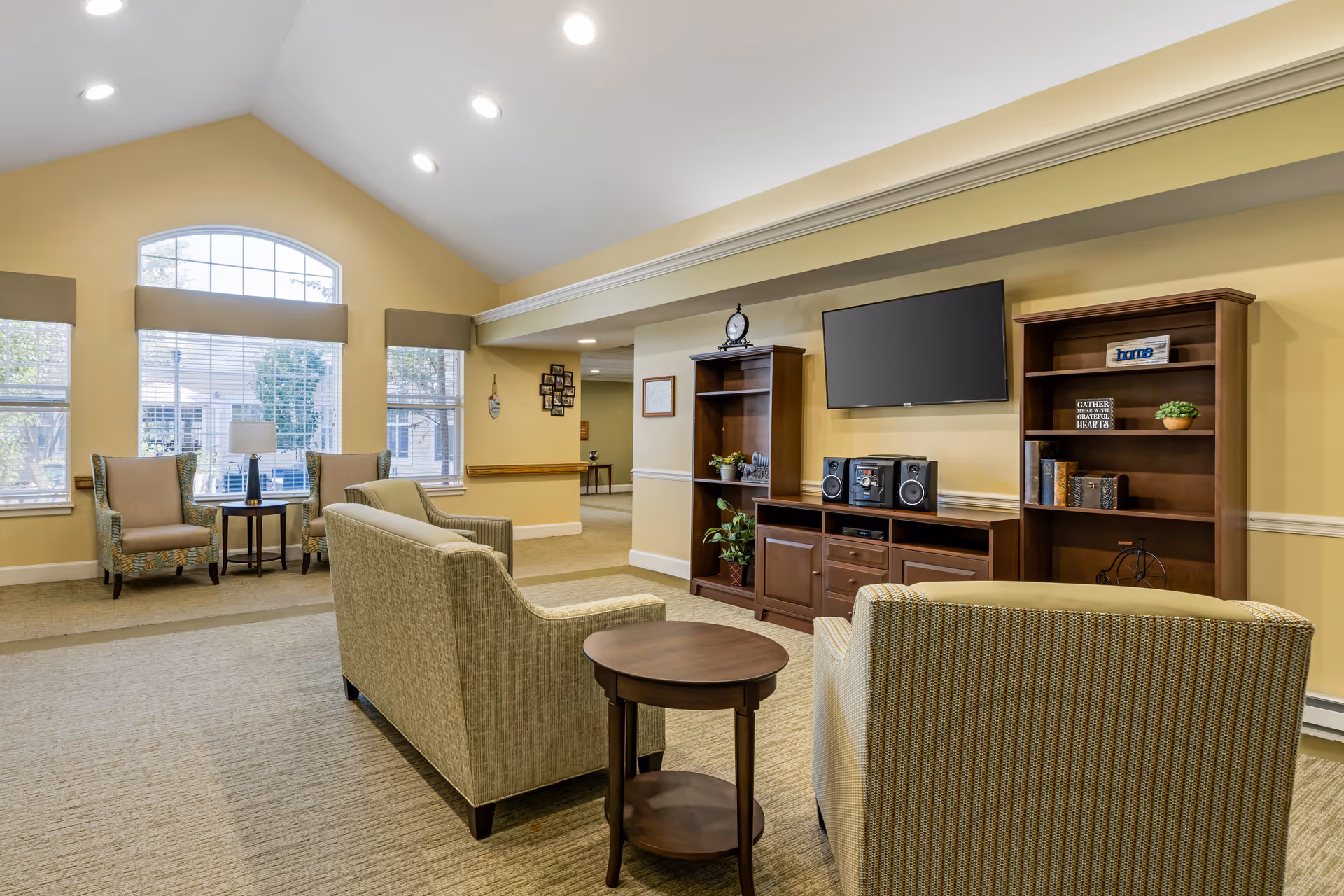 A bright and spacious living room area with beige walls and carpeted floor. The room features a large window with three sections, two armchairs with a small round table and lamp between them, a sofa, and an armchair facing a wooden entertainment center with a flat-screen TV mounted above it. The entertainment center also holds a stereo system and decorative items on the shelves.