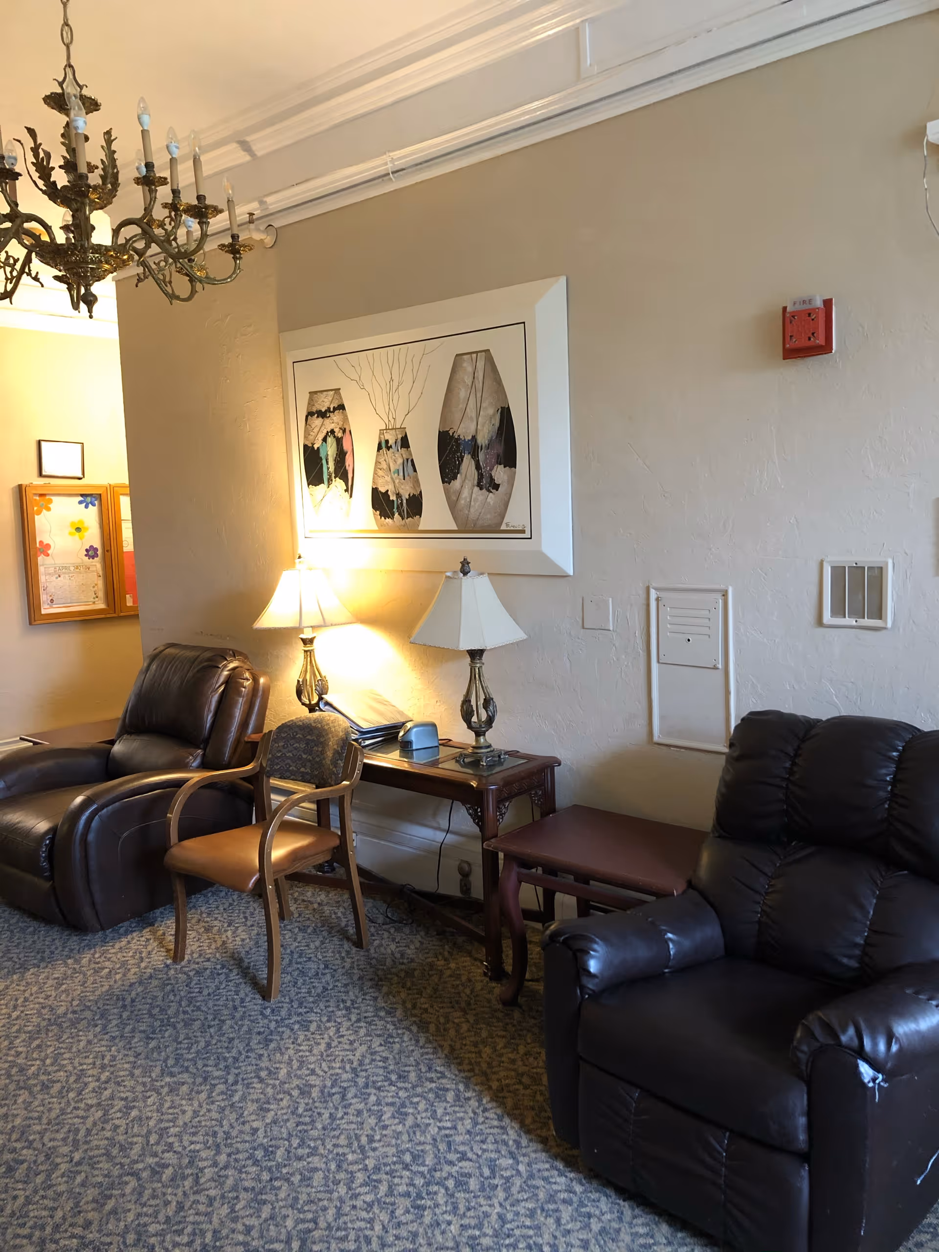 A cozy sitting area with two dark brown leather recliners and a wooden chair with cushioned seat. Between the chairs is a wooden side table with two lamps and some items on it. Above the table hangs a framed artwork depicting three abstract vases. A chandelier hangs from the ceiling, and a bulletin board is visible on the far wall.