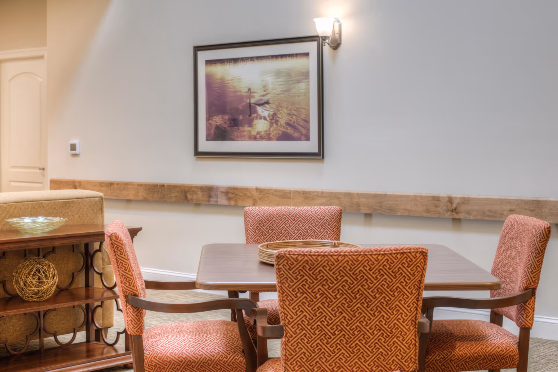 A dining area with a wooden table surrounded by four upholstered chairs with a red geometric pattern. On the table, there are two round woven placemats. Behind the table, there is a wooden console table with decorative items including a glass bowl and a spherical woven ornament. A framed picture of swans on water hangs on the wall above a wooden chair rail. A wall sconce light fixture is mounted on the wall to the right of the picture.