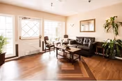 A bright living room with wooden flooring, a black leather sofa, two wooden chairs with cushions, a coffee table, a side table with a lamp, a large mirror on the wall, and a tall green plant near the corner. There are large windows and a door letting in natural light.