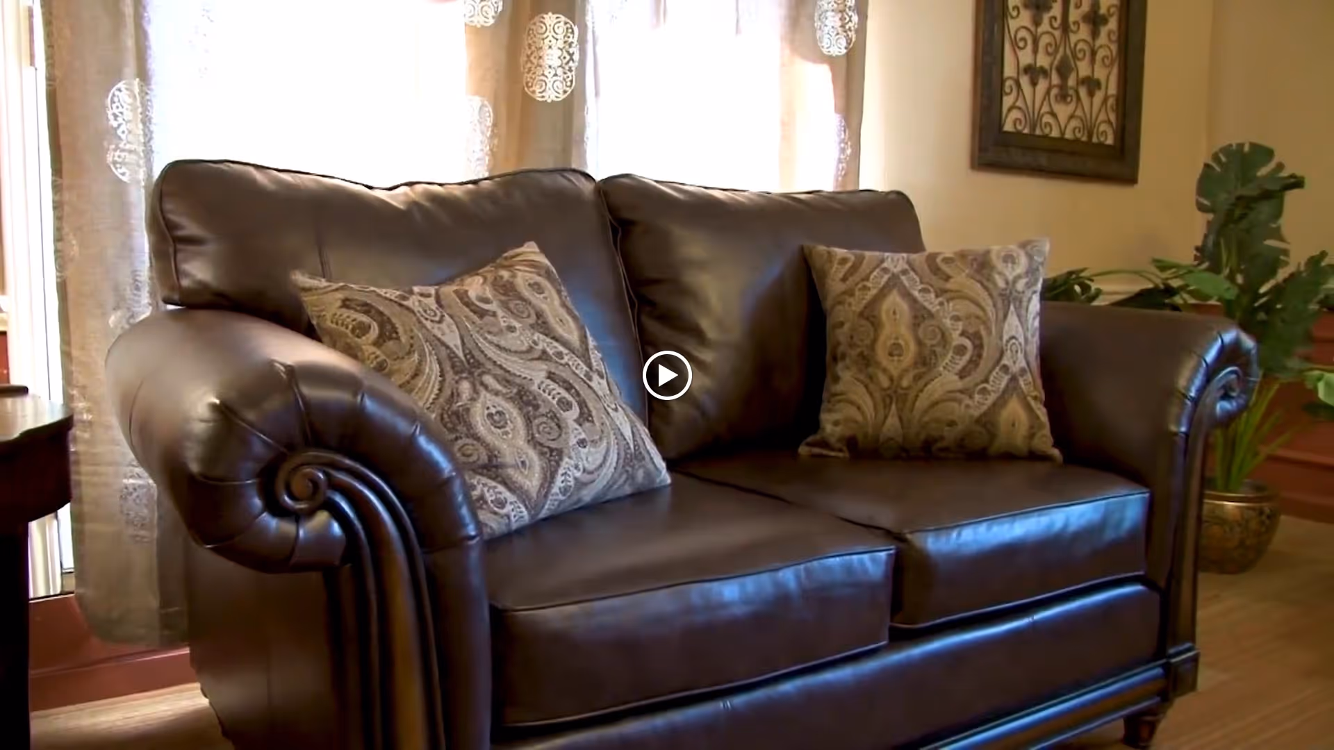 Brown leather loveseat with two patterned throw pillows in a living room with curtains, a plant, and wall art.