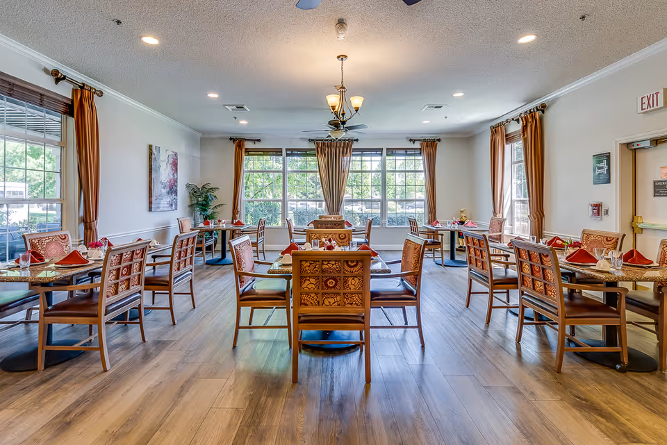 A bright and spacious dining room with multiple wooden tables and chairs arranged neatly. Each table is set with red napkins, glasses, and small flower arrangements. Large windows with brown curtains allow natural light to fill the room, and a chandelier hangs from the ceiling. The floor is wooden, and there are some decorative plants and artwork on the walls.