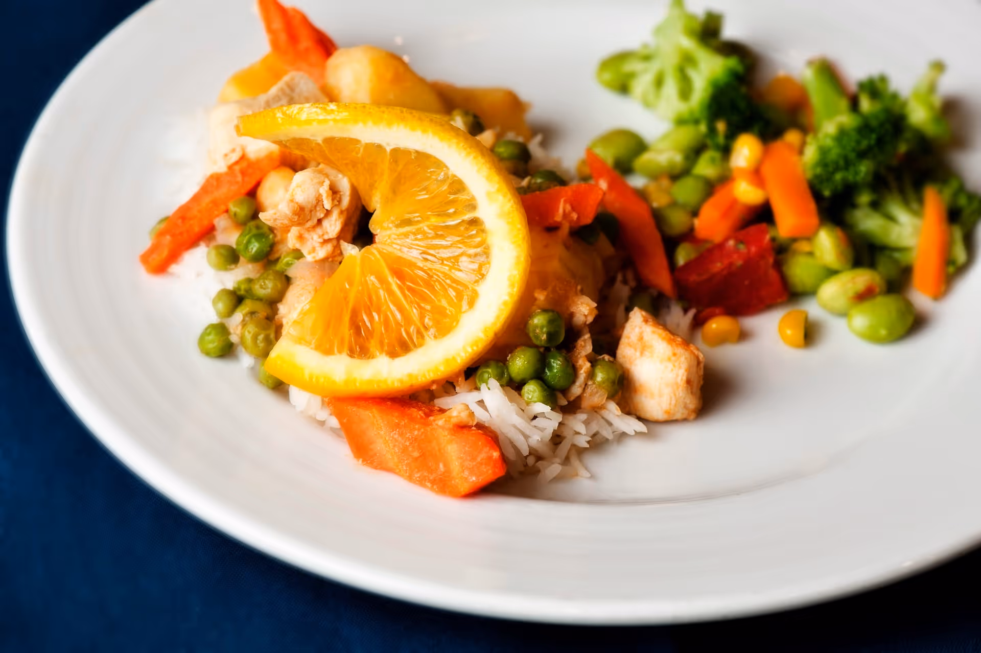 A close-up of a plate with cooked white rice topped with peas, carrots, pieces of chicken, and a slice of orange. On the side, there is a serving of mixed vegetables including broccoli, corn, edamame, and carrots.