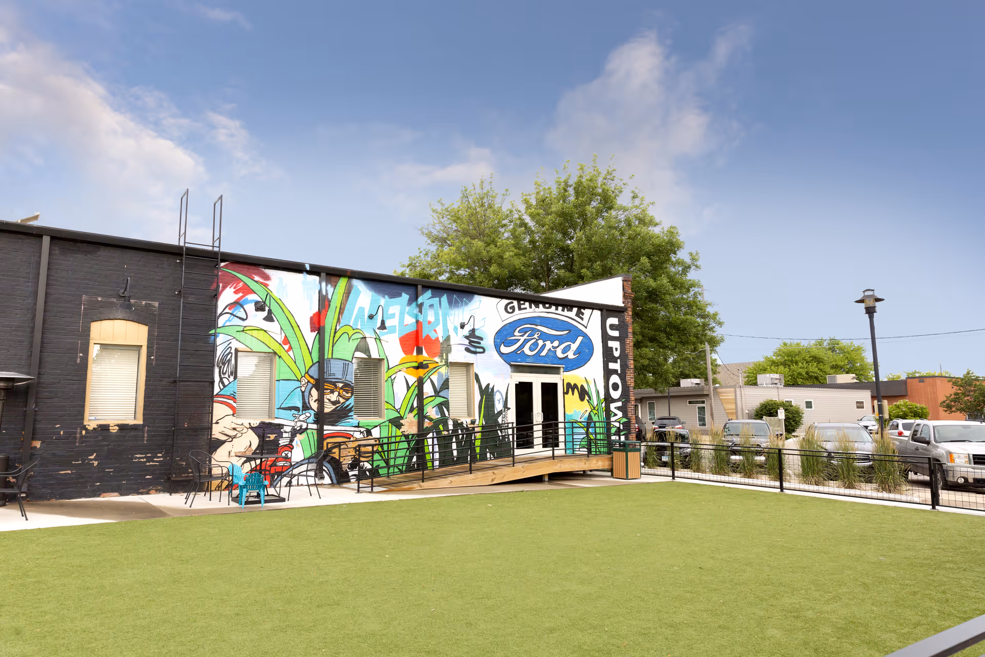 Outdoor area with a green artificial turf lawn in front of a building featuring colorful graffiti art, including a large 'Genuine Ford' logo and the word 'Uptown'. There are a few chairs and tables near the building, a ramp leading to a door, and several parked cars in the background under a partly cloudy sky.