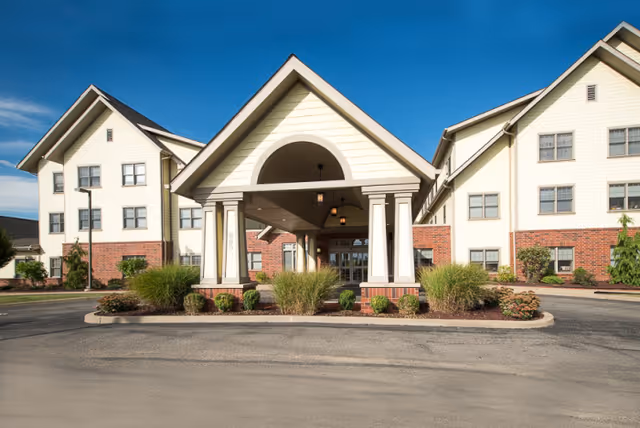 Front exterior view of a senior living facility building with a covered entrance supported by columns, surrounded by landscaping and a paved driveway under a clear blue sky.