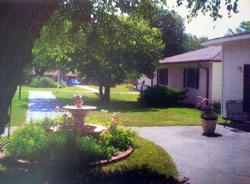 Outdoor view of a senior living facility courtyard with a paved walkway, green grass, a flower bed with a decorative fountain, trees providing shade, and single-story buildings with windows and a potted plant near the walkway.