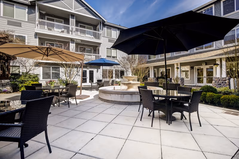Outdoor patio area at Aegis Living Shoreline with multiple tables and chairs under large umbrellas, a central stone water fountain, surrounded by a multi-story building with balconies and windows, under a clear blue sky.