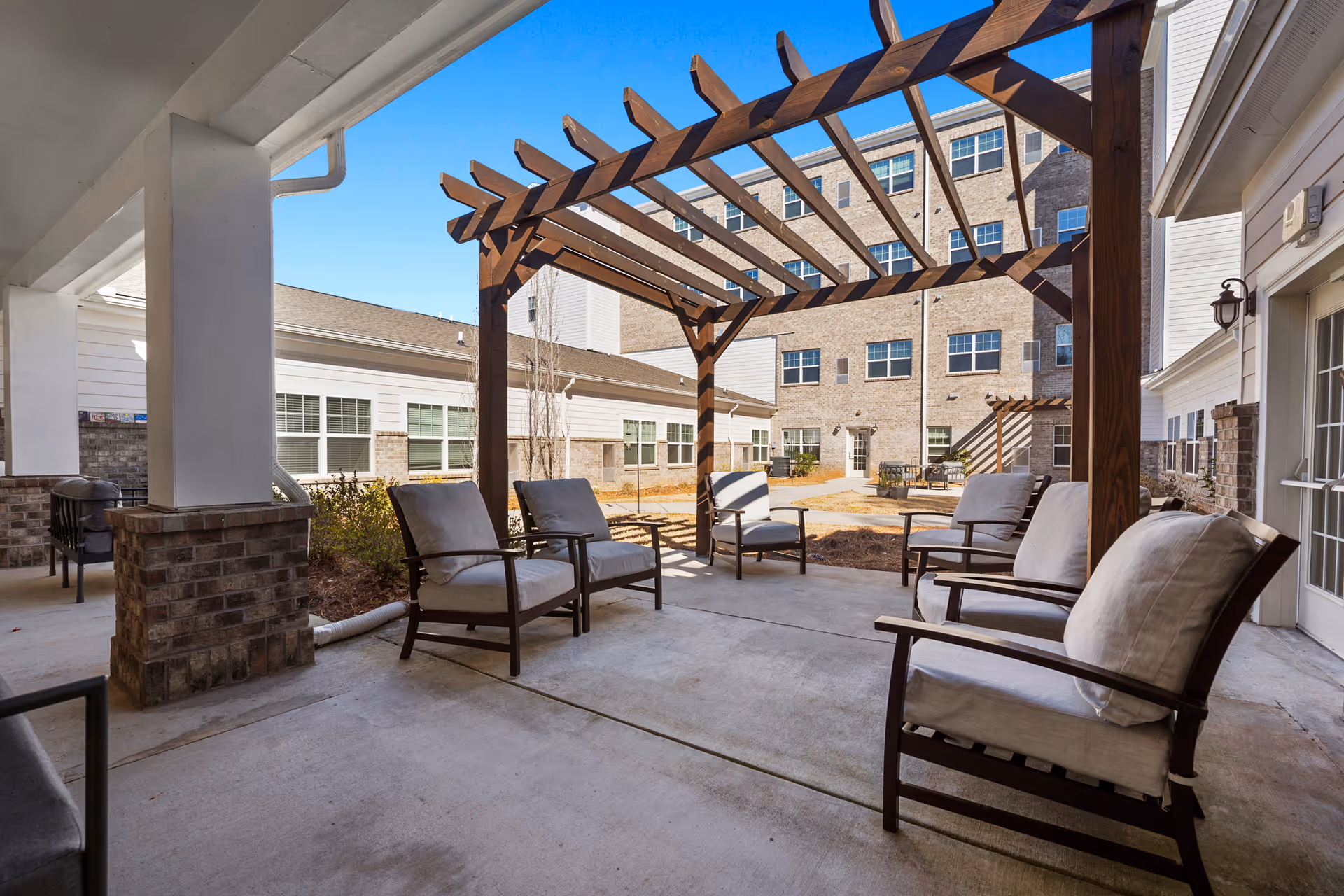 Outdoor seating area at Alto Johns Creek featuring cushioned chairs arranged under a wooden pergola with a multi-story building in the background and clear blue sky.
