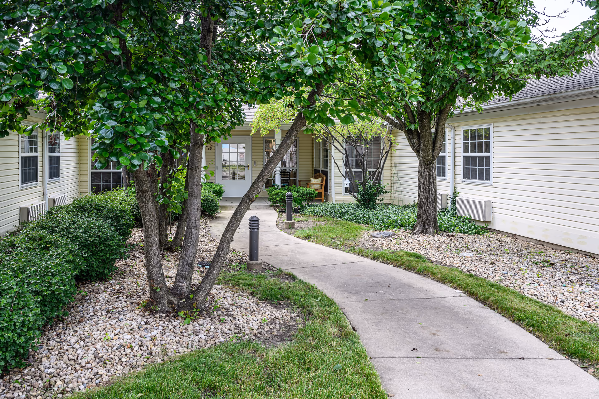 A curved concrete pathway leads through a landscaped garden area with green grass, bushes, and trees towards the entrance of a beige building with white-framed windows and a glass door. There are two black outdoor lights along the path and a wooden chair on the porch near the door.