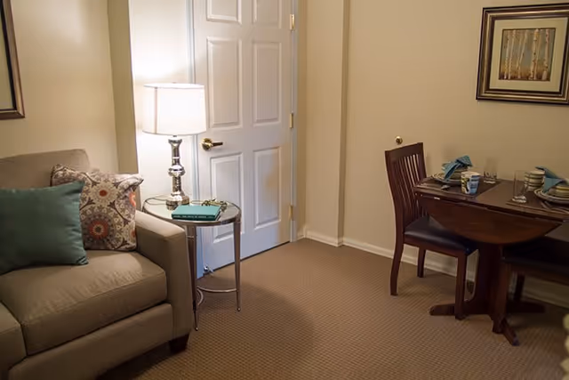 A cozy interior room featuring a beige armchair with decorative pillows, a small round side table with a lamp and a book, a white door, and a wooden dining table set with plates, glasses, and napkins against a beige wall with a framed picture.
