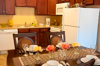 A kitchen and dining area with wooden cabinets, a white refrigerator, dishwasher, stove, and microwave. The dining table is set with plates, glasses, silverware, and decorated with small vases holding pink and yellow flowers on a patterned table runner.