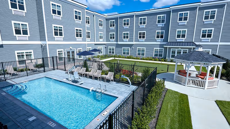 Outdoor courtyard area of The Bristal Assisted Living at Bethpage featuring a fenced swimming pool with lounge chairs, a putting green, a white gazebo with red cushioned seating, and a three-story building with multiple windows surrounding the courtyard.