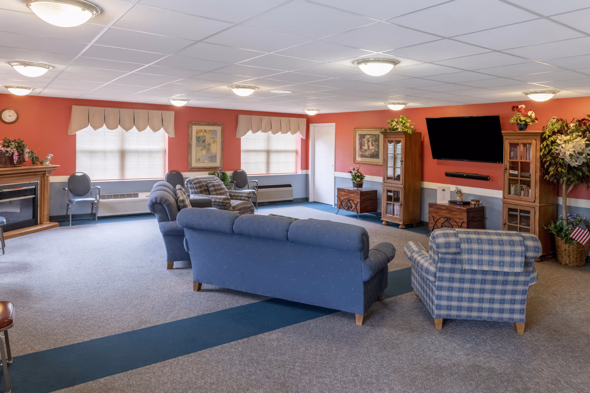 A bright communal living room with sofas and armchairs arranged around a wall-mounted TV, cabinets, and a fireplace in a senior facility.