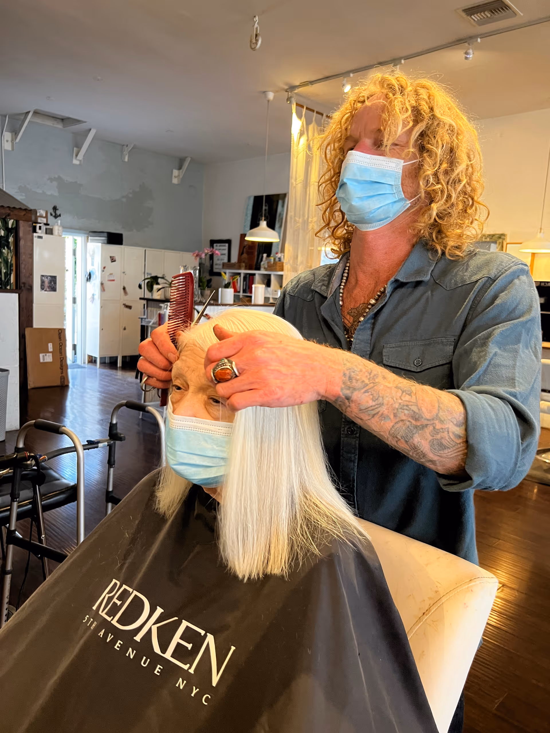 A hairstylist wearing a blue shirt and a face mask is combing the hair of an elderly woman with white hair who is also wearing a face mask and a black salon cape with the Redken logo. The setting appears to be a salon area within a senior living facility, with lockers and chairs visible in the background.