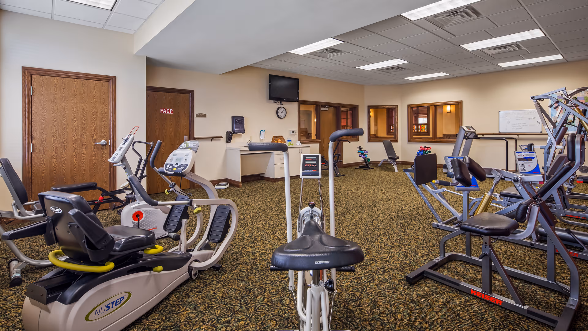 Interior view of a fitness room in a senior living facility with various exercise equipment including stationary bikes, weight machines, and benches. The room has patterned carpet, beige walls, wooden doors, a wall-mounted TV, and a clock.
