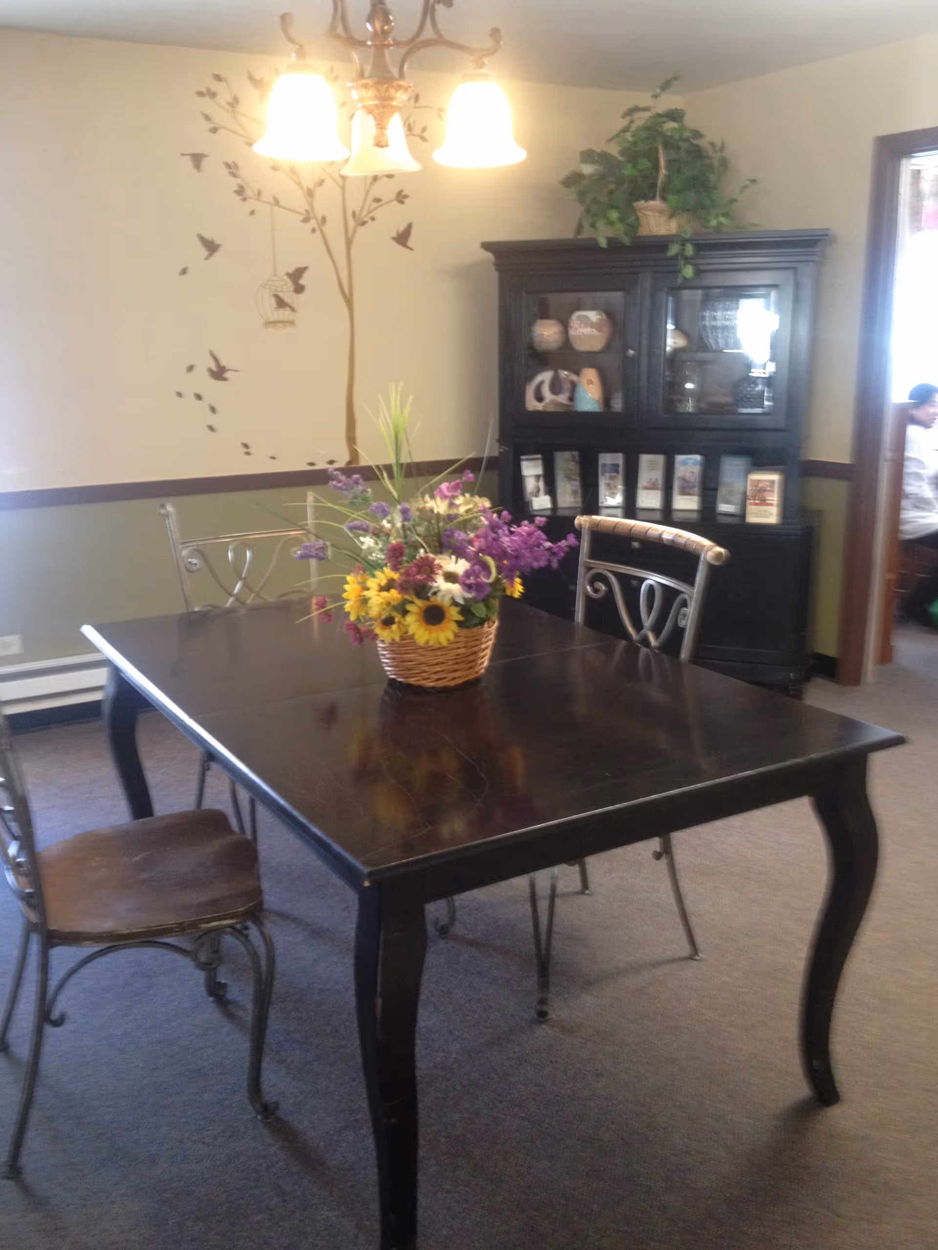 Dining room with a dark wooden table and a basket of flowers, metal chairs, a display hutch, and a bird/tree wall decal.