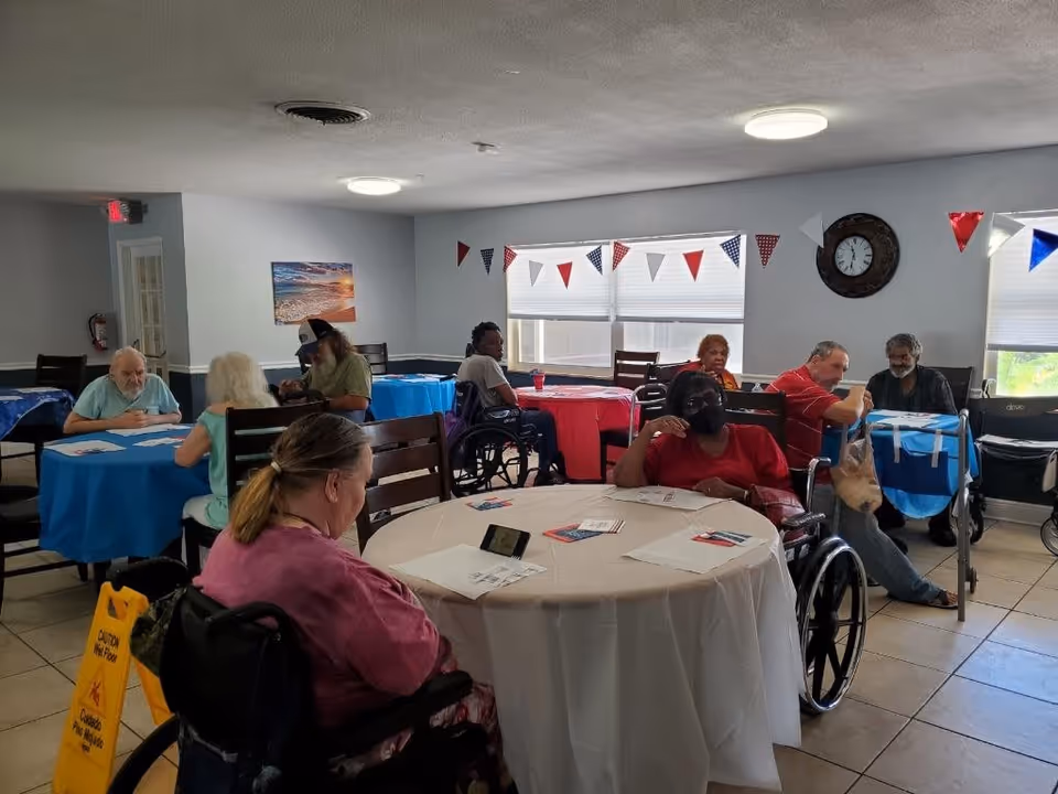 A group of elderly people, some in wheelchairs, sitting around tables covered with red, white, and blue tablecloths in a common room decorated with red, white, and blue triangular pennant banners. The room has tiled floors, a clock on the wall, and windows with blinds partially drawn.