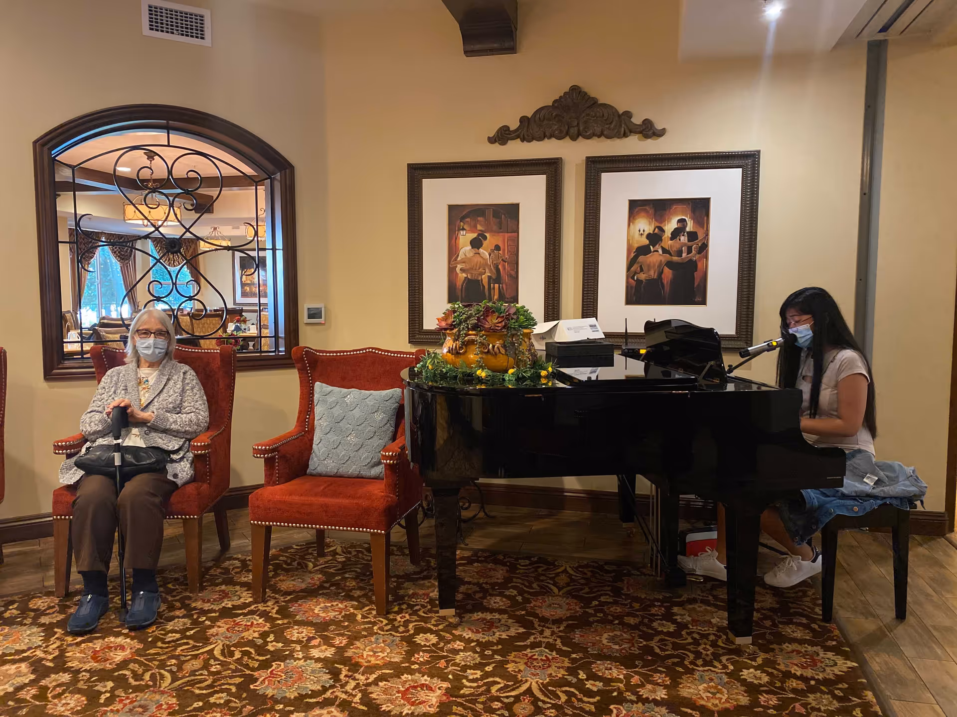 A cozy lounge with a grand piano, framed artwork, and two masked women—one seated in a red armchair and one playing the piano.