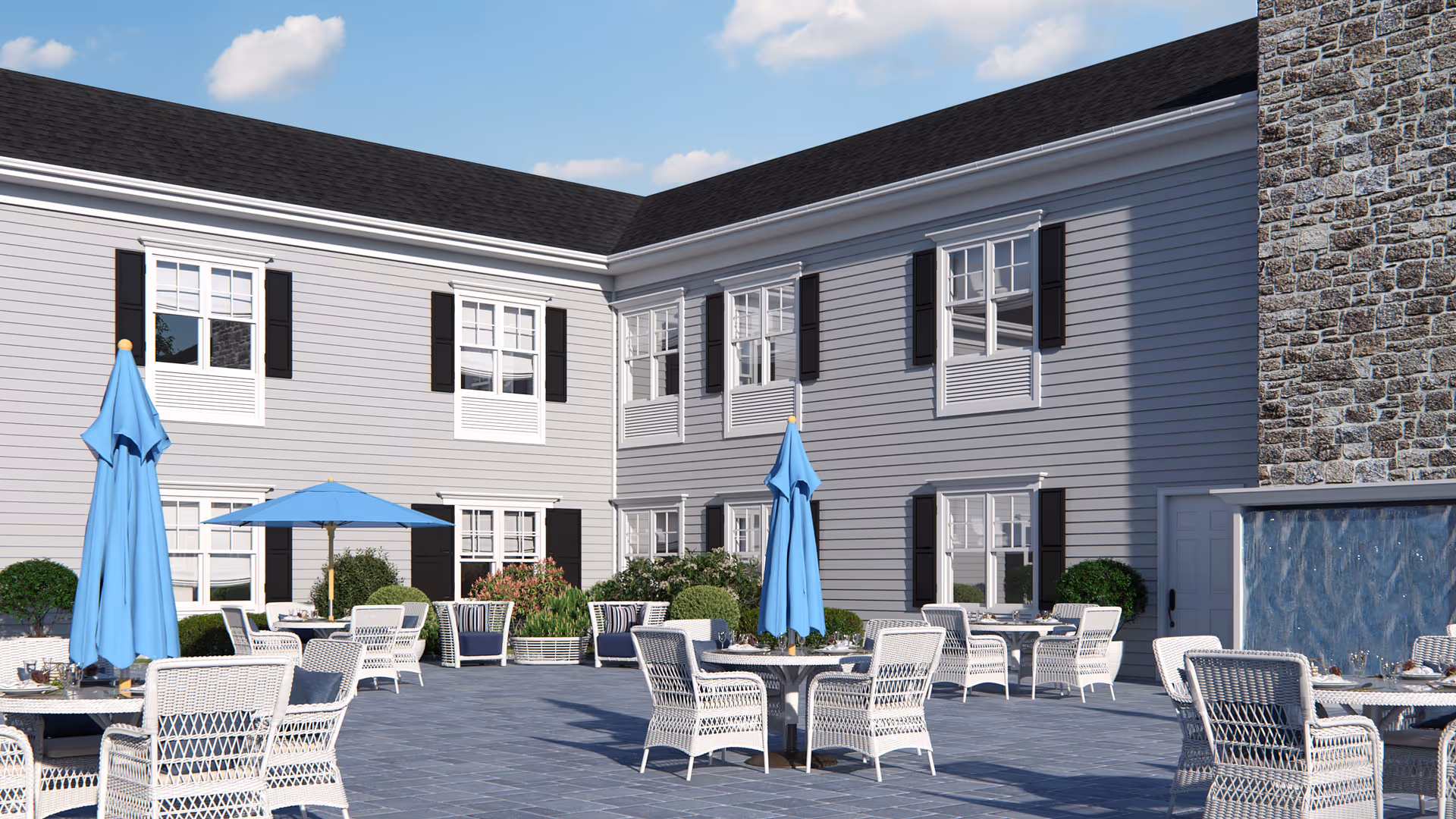 Outdoor courtyard area of The Bristal Assisted Living at Somerset featuring multiple white wicker tables and chairs with blue umbrellas, surrounded by a two-story building with gray siding, white-framed windows, and black shutters. There are plants and shrubs along the building walls and a stone wall with a water feature on the right side.