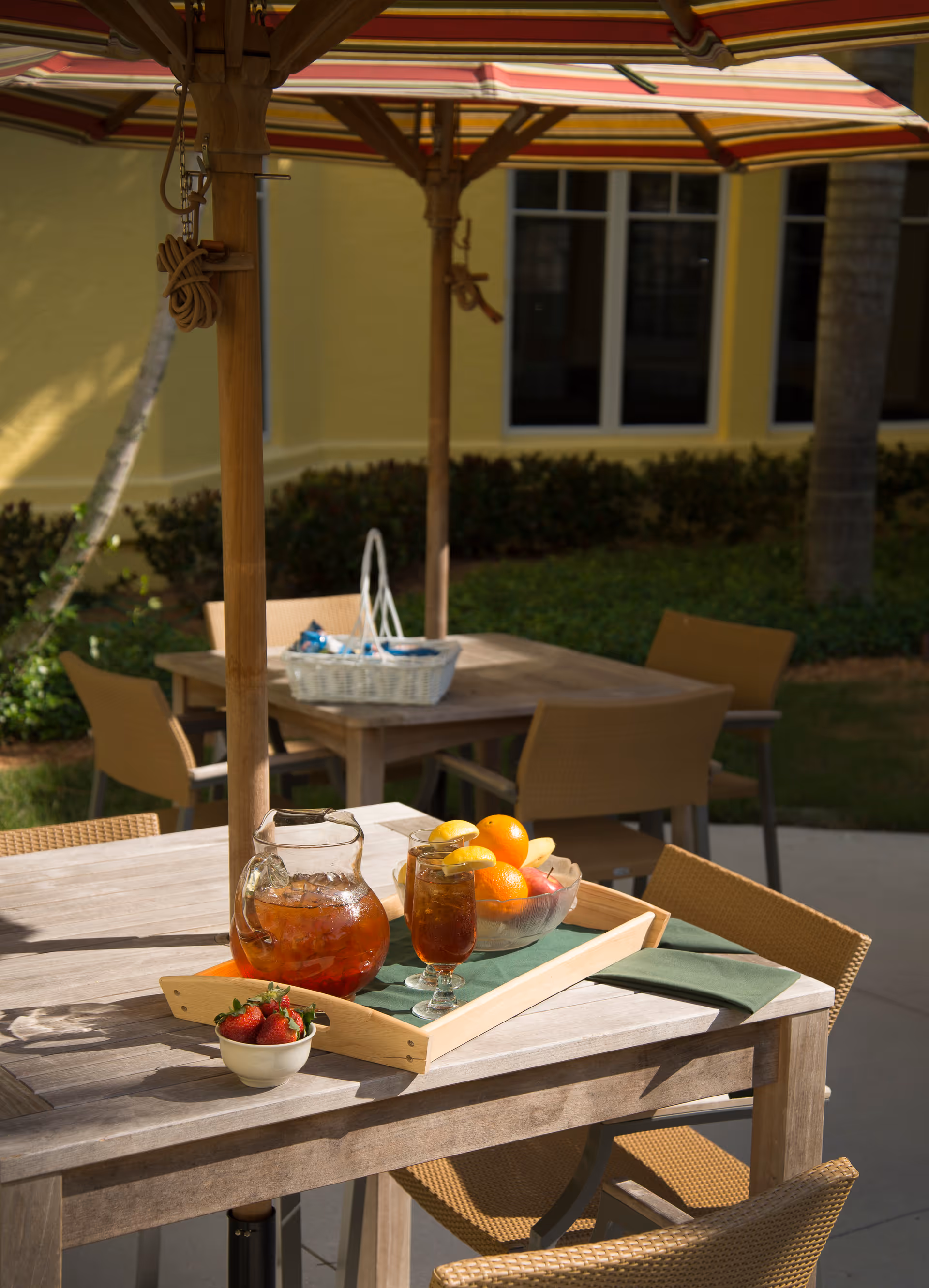 Patio dining table under a striped umbrella with a pitcher and glass of iced tea, a bowl of fruit, and surrounding chairs.