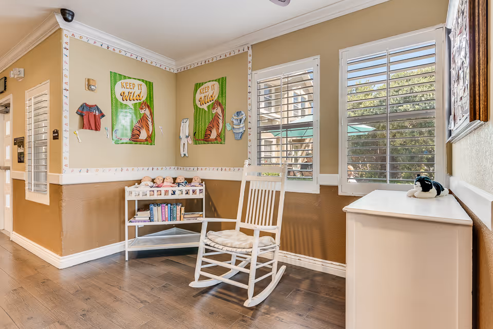 A cozy corner in a senior living facility with a white rocking chair, a small white shelf holding dolls and books, and two windows with white shutters letting in natural light. The walls are painted beige and brown with a decorative border featuring small animal illustrations. Two green posters with a tiger and the text 'KEEP IT WILD' are displayed on the wall.