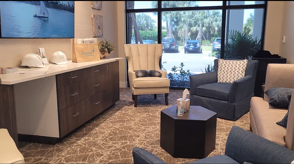 A cozy living room area with a beige armchair, a blue armchair, and a beige sofa with patterned pillows. A dark hexagonal coffee table in the center holds tissues and hand sanitizer. A large window shows a view of parked cars and greenery outside. A wooden cabinet with white countertop holds two white helmets, a paper bag labeled 'Inspired Living,' and some decorative items. A TV mounted on the wall displays a sailboat on water.