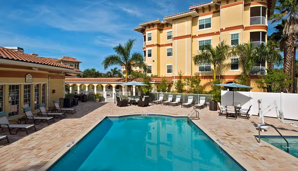 Outdoor swimming pool area at Sandalwood Village with lounge chairs, tables with umbrellas, palm trees, and a multi-story yellow building in the background under a clear blue sky.