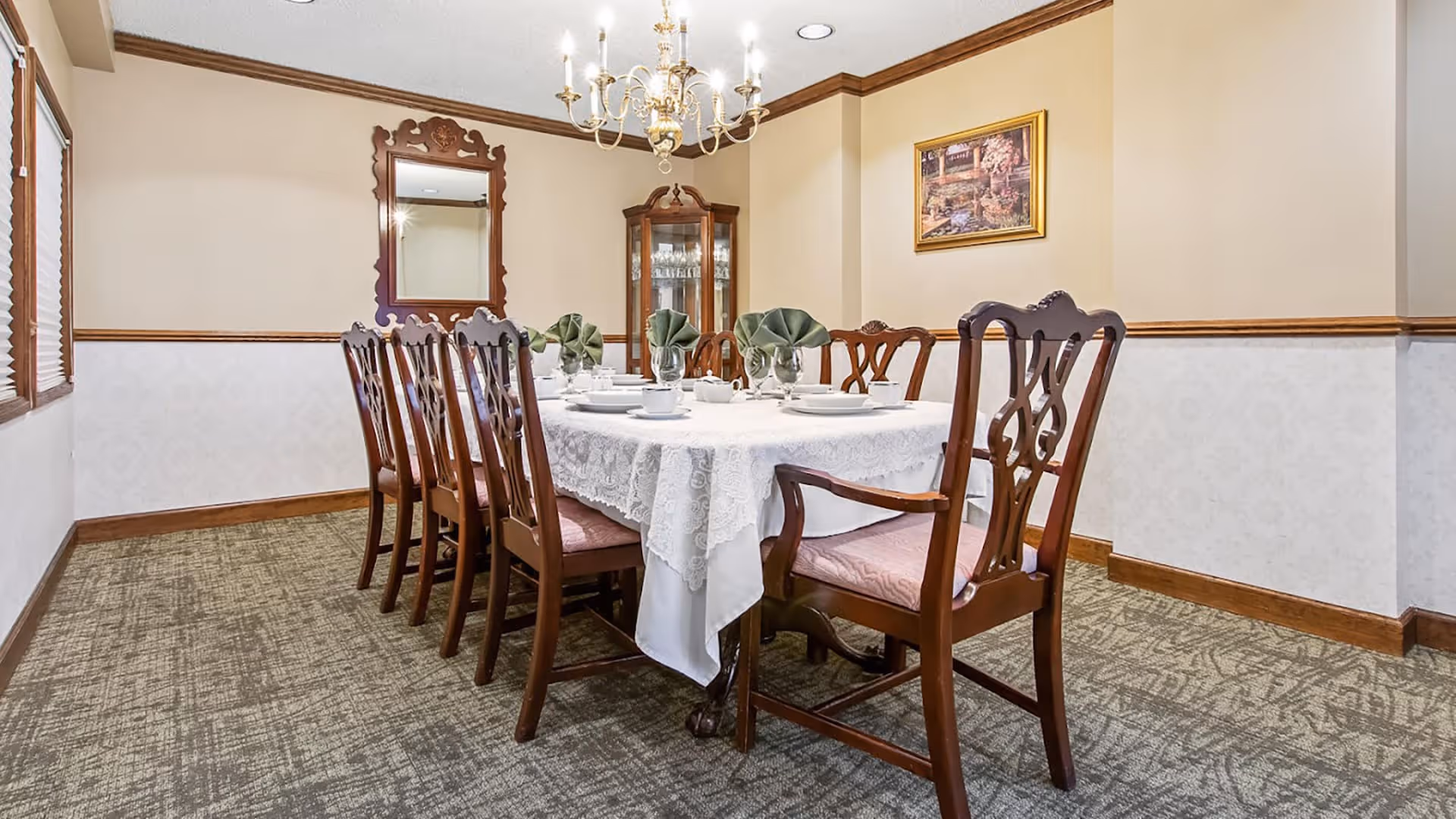 A formal dining room with a long rectangular table covered with a white lace tablecloth. The table is set with white plates, cups, and green folded napkins. There are eight wooden chairs with cushioned seats around the table. The room has beige walls with wooden trim, a chandelier hanging from the ceiling, a large decorative mirror on one wall, a framed painting on another, and a glass-fronted cabinet in the corner.