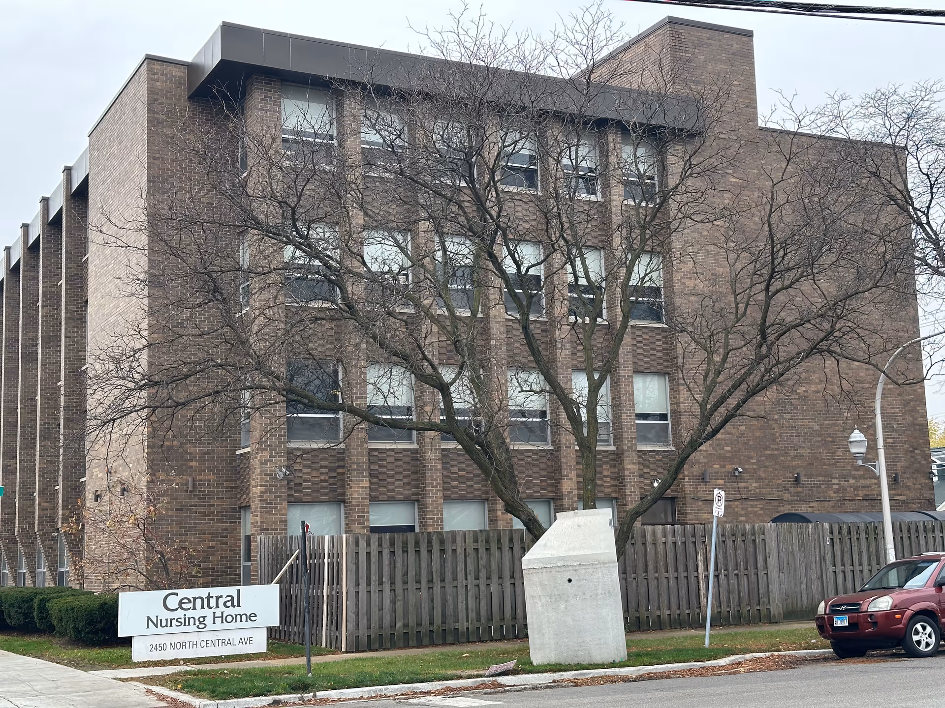 Exterior view of a three-story brick building with multiple windows, a leafless tree in front, a wooden fence, and a sign that reads 'Central Nursing Home, 2450 North Central Ave'. A red SUV is parked on the street nearby.