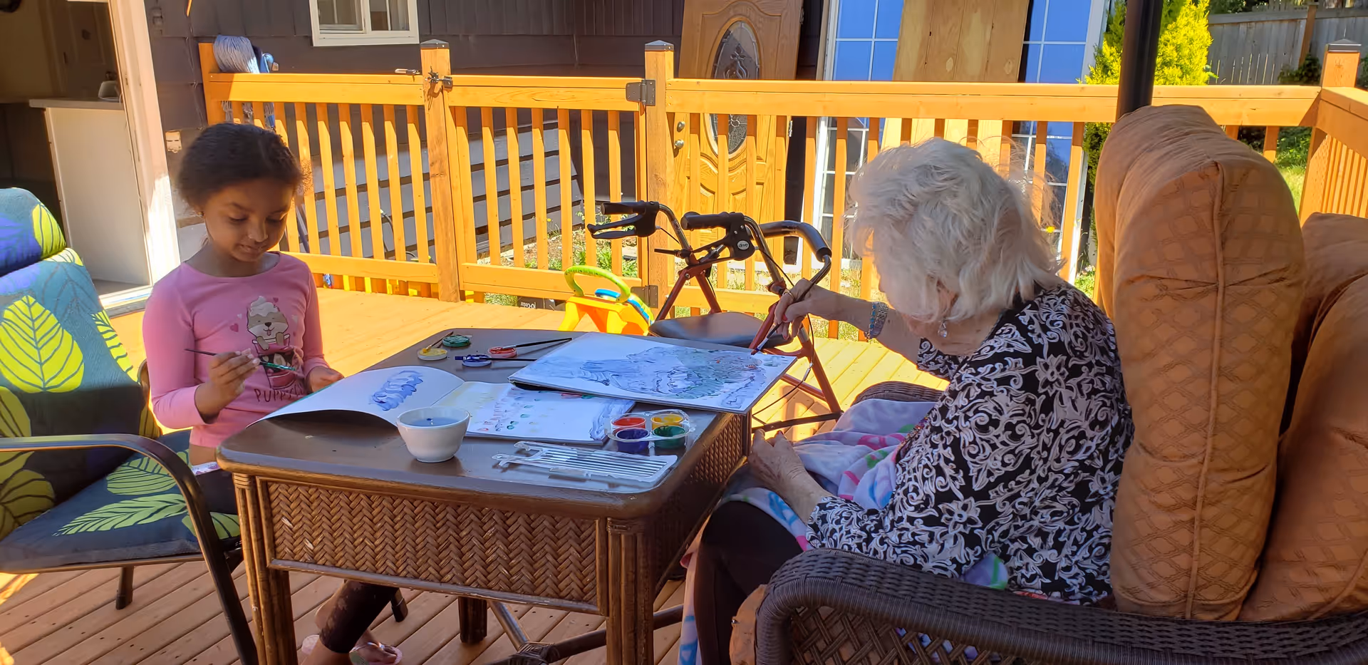 An elderly woman and a young girl painting together at a table on a sunny wooden deck with patio furniture and a walker nearby.