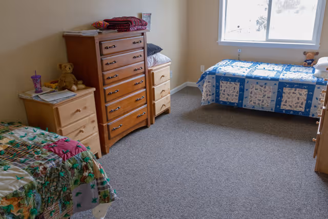A simple bedroom with two twin beds, each covered with colorful patchwork quilts. Between the beds are wooden dressers and nightstands, with a teddy bear and a cup on one of the nightstands. A window on the far wall lets in natural light, illuminating the carpeted floor and beige walls.