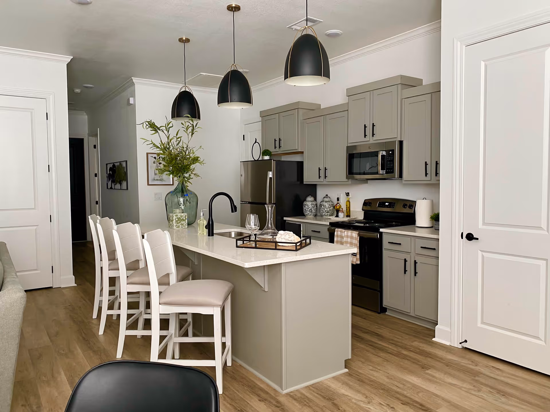 Modern kitchen with light gray cabinets, black appliances including a stove and microwave, and a large island with a white countertop. The island has a built-in sink with a black faucet and four white cushioned bar stools. Three black pendant lights hang above the island. There is a large green vase with branches on the island and decorative items on the countertop. The floor is light wood, and white walls and doors frame the space.