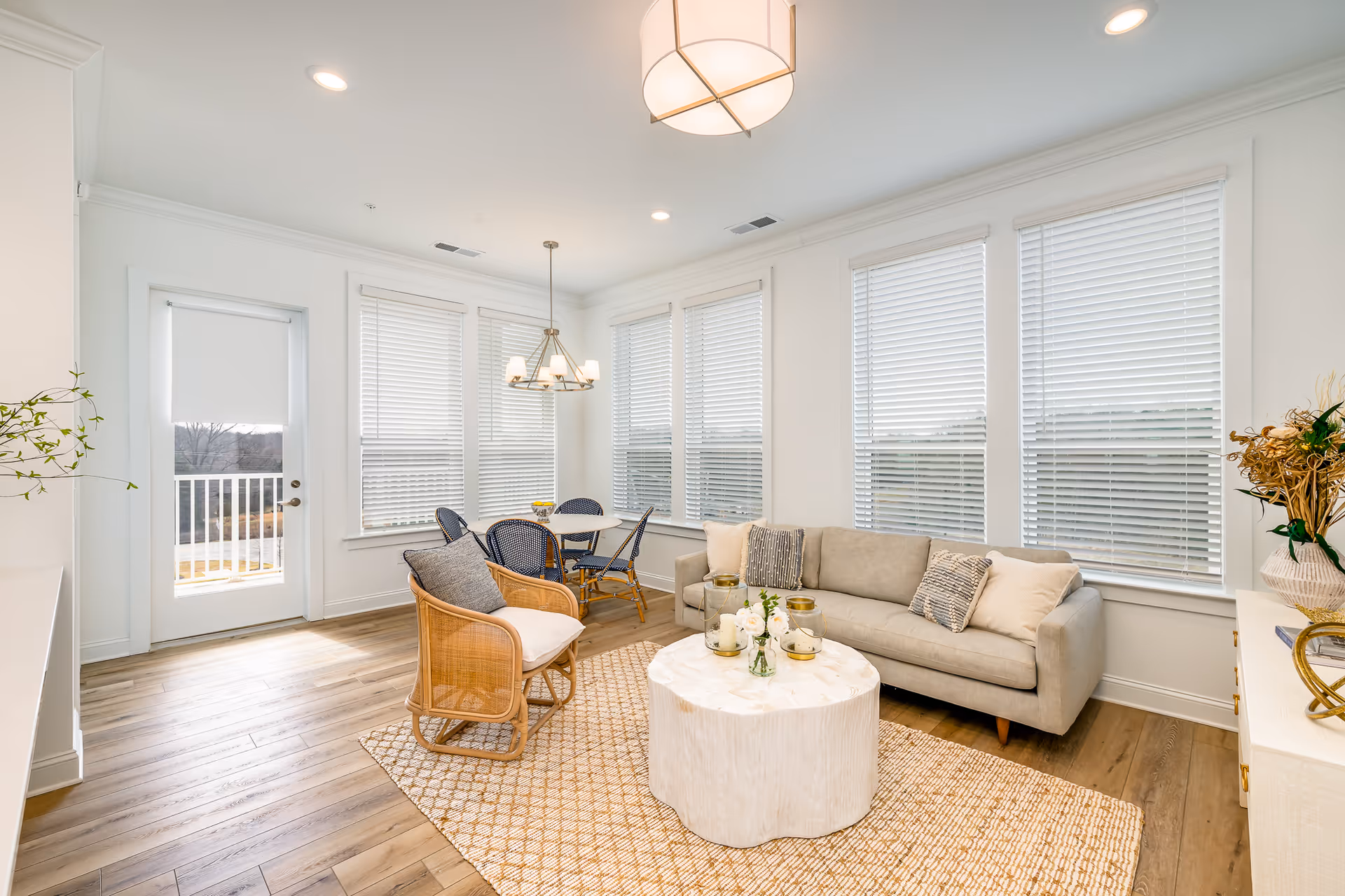 Bright and airy living room area with a beige sofa adorned with cushions, a round white coffee table with decorative items, a woven armchair, and a dining table with four chairs near large windows with white blinds. The room has light wood flooring and white walls, with a door leading outside.
