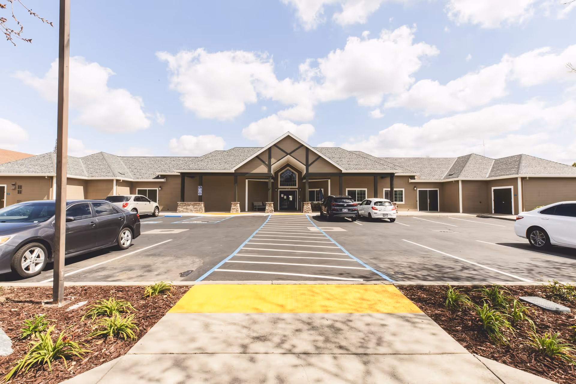 Front exterior view of a single-story building with a peaked roof and covered entrance, surrounded by a parking lot with several parked cars and a sidewalk with a yellow tactile paving strip leading to the entrance under a partly cloudy sky.