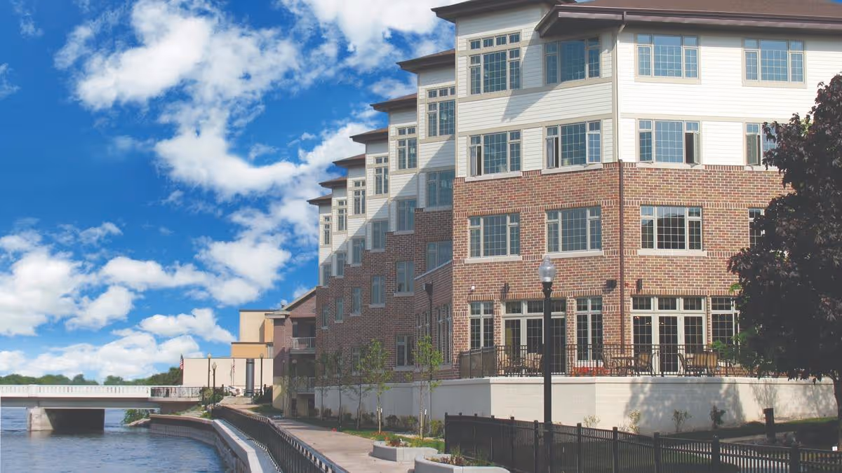 Exterior view of a multi-story senior living facility building with brick and light-colored siding, large windows, a patio area with outdoor seating, a walkway along a waterway, and a bridge in the background under a partly cloudy blue sky.
